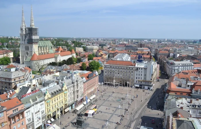 Panoramic aerial photo of Zagreb’s Ban Jelačić Square, the city’s bustling central plaza, colorful historic buildings, tram tracks, and Zagreb Cathedral’s twin spires in the distance.