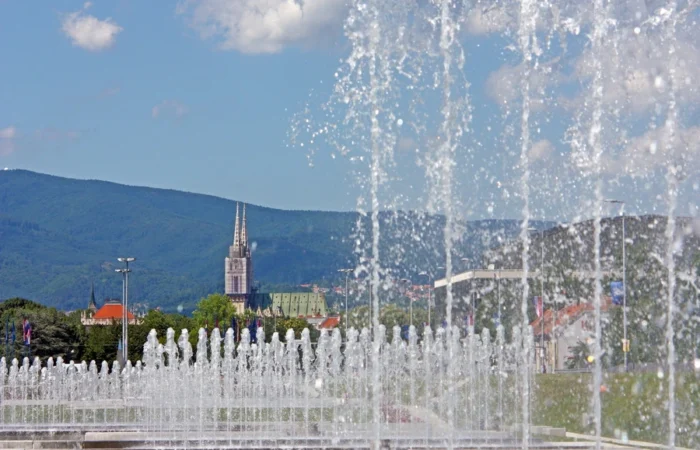 Water fountains sparkling in the foreground, with Zagreb Cathedral’s twin spires and green Medvednica mountain under a sunny sky in the distance.