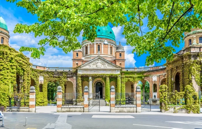 Grand neo-Renaissance arcades, domes, and lush ivy-covered walls of Mirogoj Cemetery in Zagreb, Croatia, framed by vibrant green spring leaves on a bright, clear day.