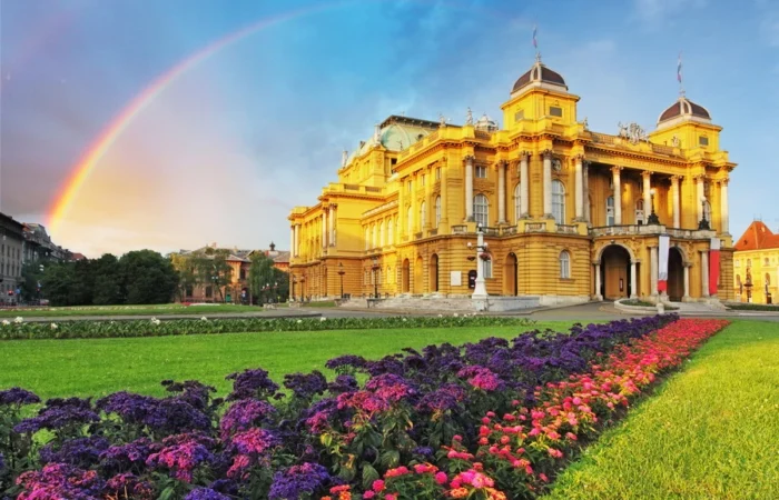 The Croatian National Theatre in Zagreb glowing in golden sunlight with a vibrant rainbow overhead, lush green lawn, and gardens of purple and red flowers in the foreground.