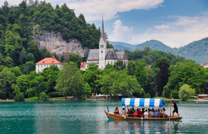 Traditional pletna boat with tourists on Lake Bled, Slovenia, with a white church and forested hills in the background.