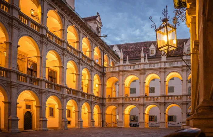 Evening view of the illuminated Renaissance arcaded courtyard of the Landhaus in Graz, Austria, with glowing arches and hanging lanterns.
