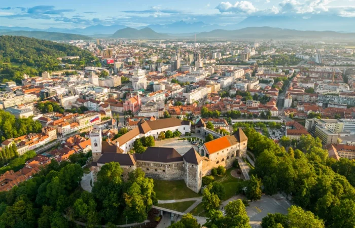 Aerial view of Ljubljana Castle surrounded by trees with the city of Ljubljana and distant mountains in the background on a sunny day.