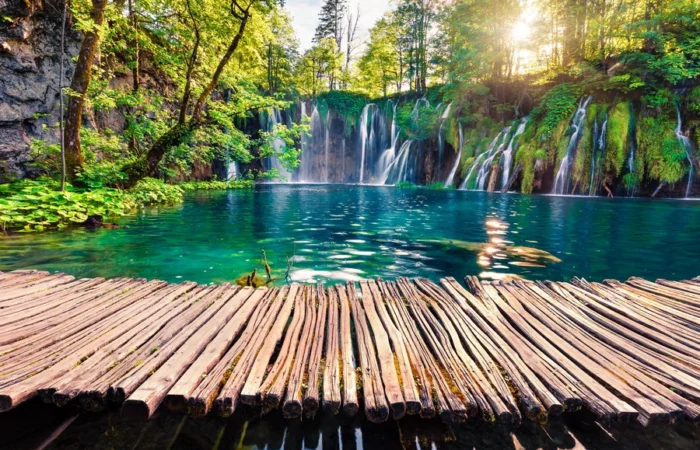 Sunlit waterfalls cascade into a turquoise lake at Plitvice Lakes National Park in Croatia, with a rustic wooden boardwalk in the foreground.