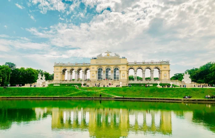 The Gloriette at Schönbrunn Palace in Vienna reflected in a green pond, with people walking along the grassy terrace under a partly cloudy sky.