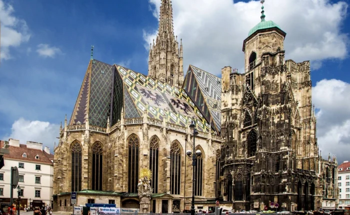 St. Stephen’s Cathedral in Vienna with its colorful tiled roof, Gothic towers, and surrounding city buildings under a bright, partly cloudy sky.
