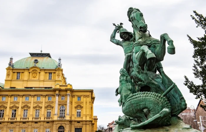 Green bronze statue of a knight on horseback slaying a dragon in front of the yellow Croatian National Theatre building in Zagreb.